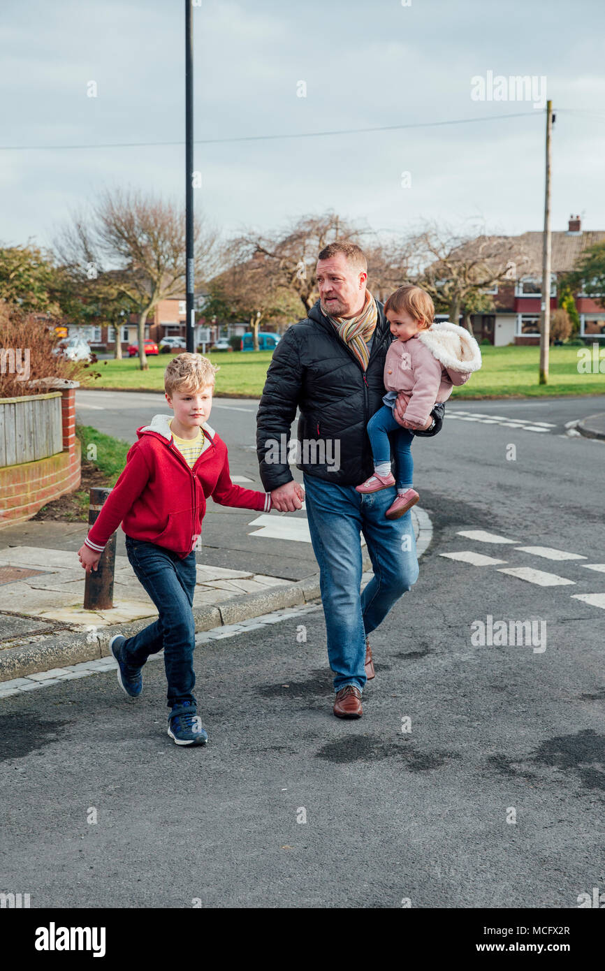 Uomo maturo è attraversare la strada con i suoi figli e il suo figlio è la mano e sta portando la sua figlia sul suo fianco. Foto Stock