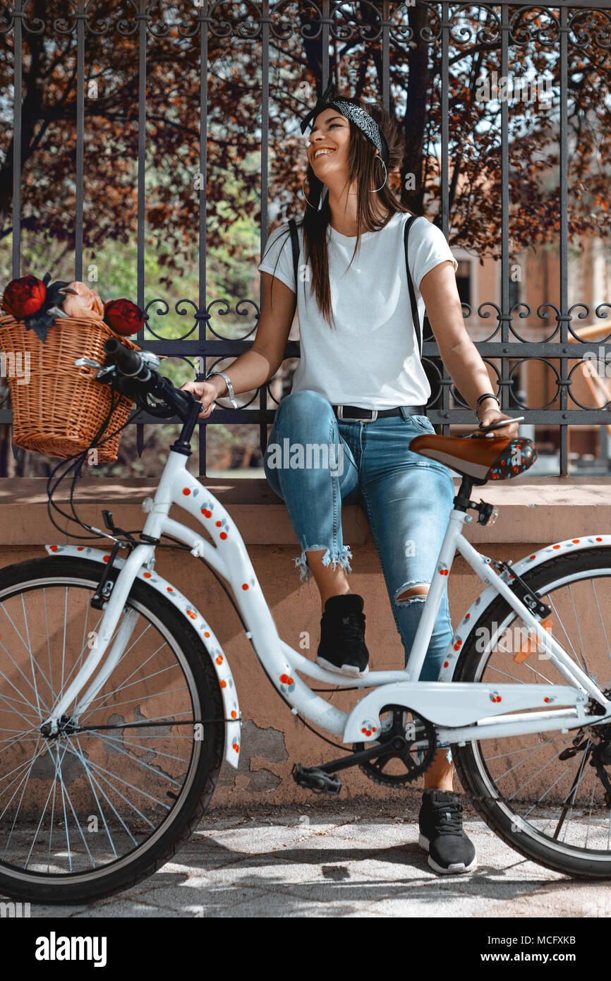 Sorridenti giovane donna sta godendo in estate il giorno soleggiato, seduto sulla recinzione della città, accanto alla moto con cesto fiorito. Foto Stock