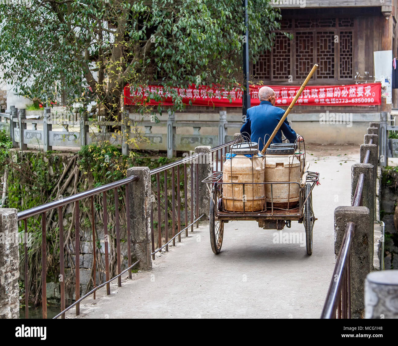 Un anziano uomo cinese che trasportano grandi vasche sul retro di un triciclo attraversa un ponte in Daxu, Cina. Egli porta anche il bambù stanghe o forcelle. Foto Stock