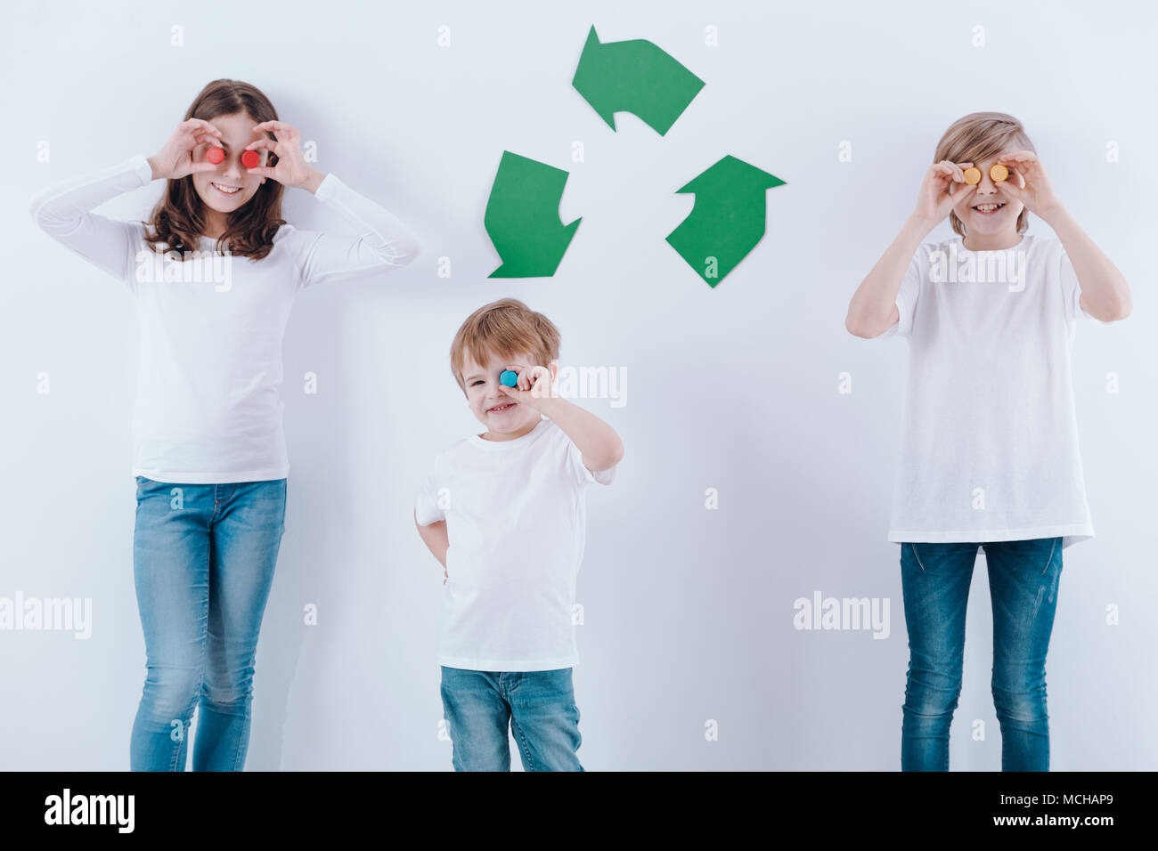 Dei bambini felici con tappi colorati contro il muro bianco verde con il simbolo di riciclaggio Foto Stock