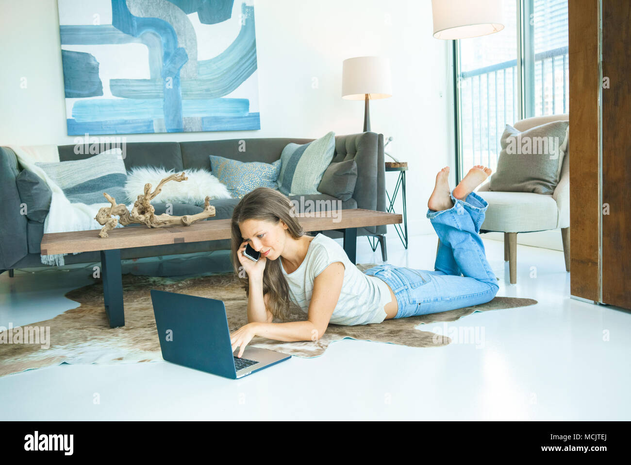 Giovane donna utilizzando laptop e parlando al telefono mentre è sdraiato sul tappetino a casa Foto Stock
