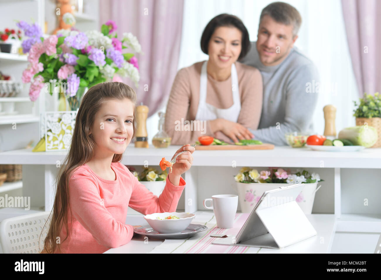 Ragazza sorridente mangiare e utilizzo di tablet in cucina con i genitori Foto Stock