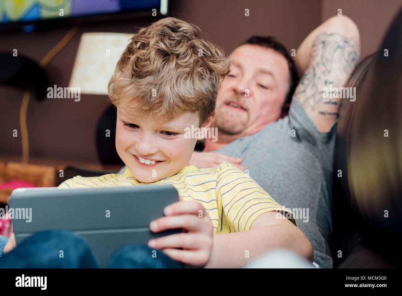 Little Boy è seduta sul suo papà in ginocchio sul divano di casa, utilizzando una tavoletta digitale. Il suo papà è lui a guardare. Foto Stock