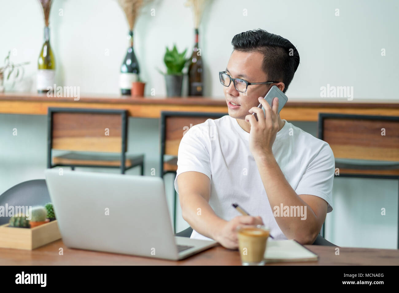 Casual Uomo asiatico parlando al telefono cellulare funziona con computer portatile sul tavolo di legno nella caffetteria,moderno stile di vita digitale concetto.influencer blogger Foto Stock