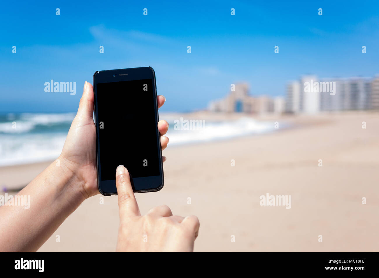 Donna con le mani in mano utilizza lo smartphone sulla spiaggia di toccare lo schermo con le onde, profondo cielo blu e sabbia dorata in background. Schermo nero sul telefono. Foto Stock