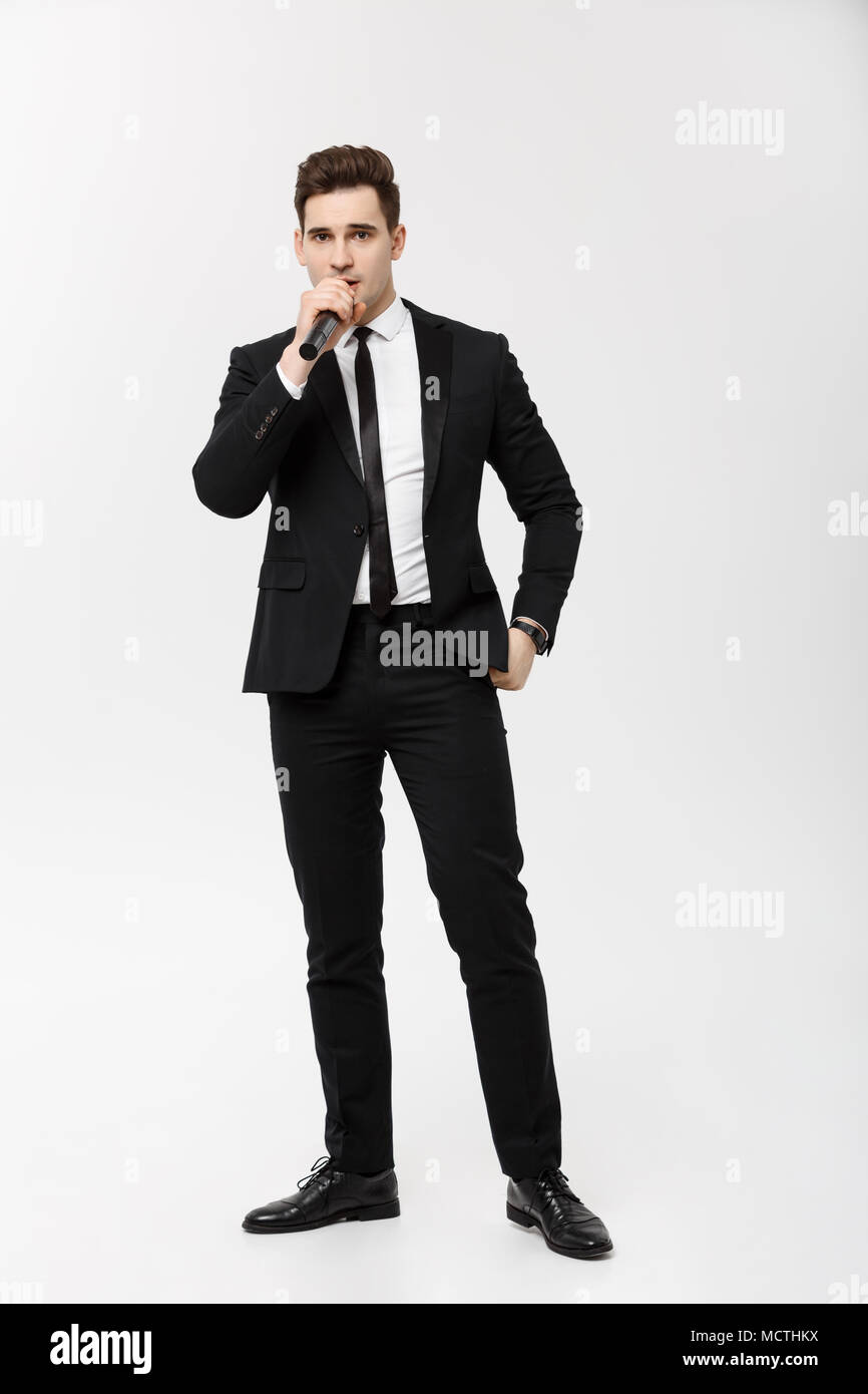 Il concetto di business: ritratto a figura intera giovane uomo in abito nero è in possesso di un microfono e di canto e posa contro uno sfondo bianco Foto Stock