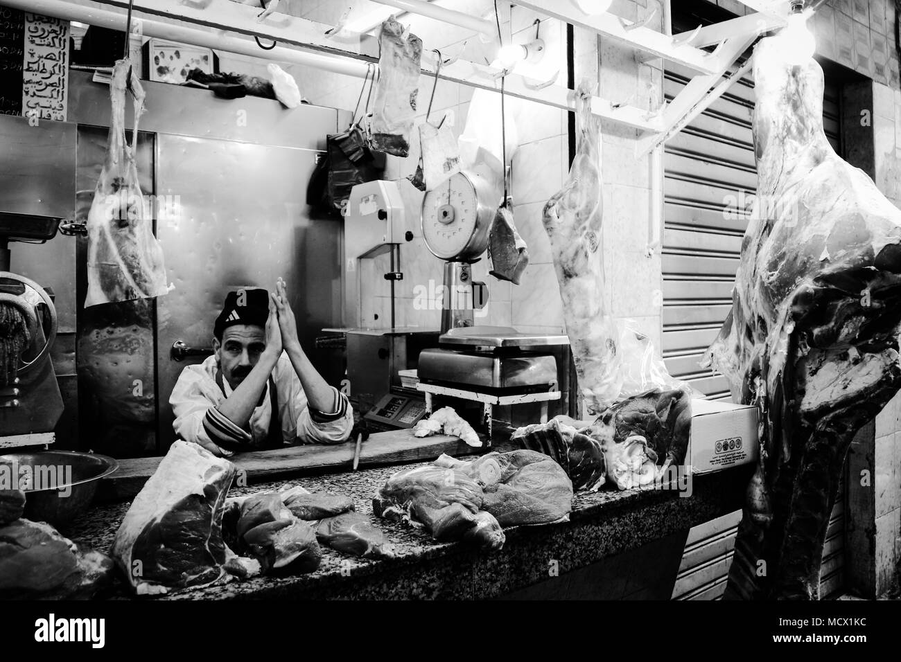 Ritratto in bianco e nero di un macellaio seduto tra carne appesi dietro il bancone della sua macelleria presso il vecchio souk di Casablanca, Marocco Foto Stock