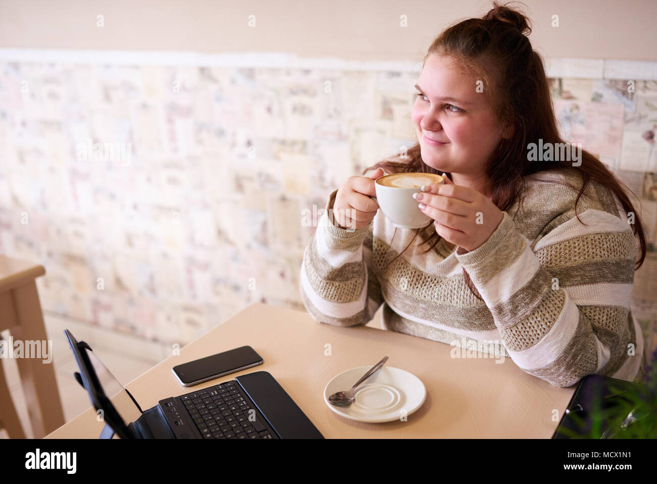 Giovane femmina adulta donna sorridente mentre guardando al di fuori della fotocamera e tenendo le sue mattina caffè con entrambe le mani, pronto per iniziare il lavoro una volta ha la sua dose giornaliera di caffeina. Foto Stock