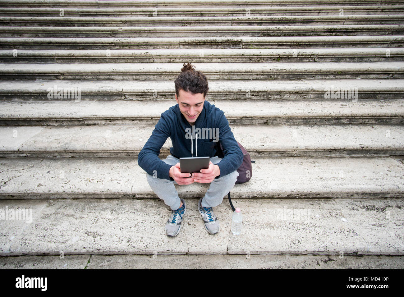 Bel giovane in abbigliamento sportivo con capelli ricci seduti sulle scale e lavorando su tablet Foto Stock