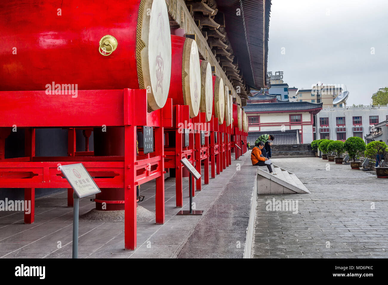 Una fila di grandi rossi tradizionali tamburi cinesi montata nella torre del tamburo, Xi'an, Cina. Una donna si siede su alcuni gradini in pietra la lettura di un libro guida. Foto Stock