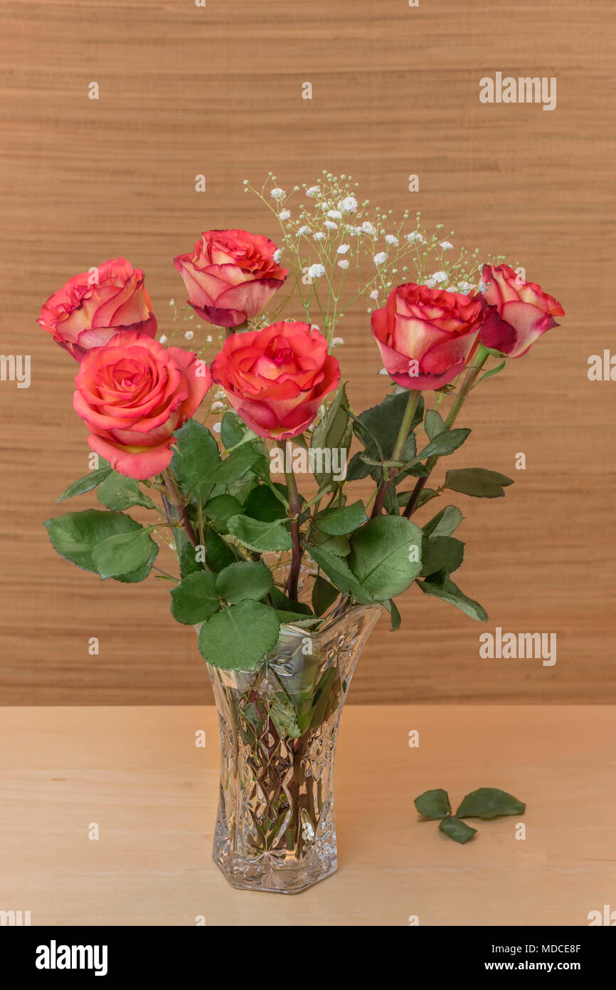 Rose rosse con foglie verdi in un vaso di vetro con acqua riposare su una tavola di legno, sfondo marrone e bianco sfocato piccoli fiori Foto Stock