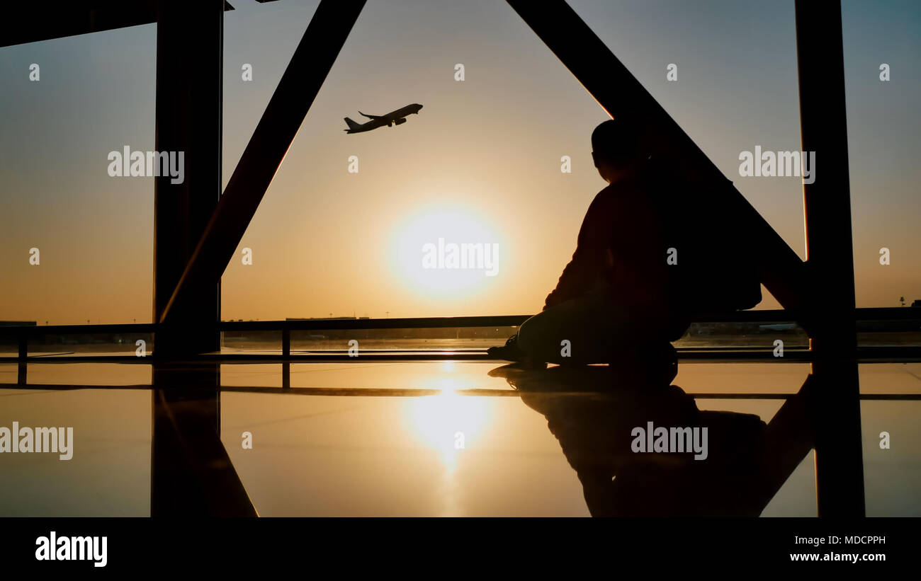 Silhouette di un turista guy guardando il decollo del piano seduta alla finestra di airport al tramonto di sera. Concetto di viaggio, la gente in aeroporto. Foto Stock