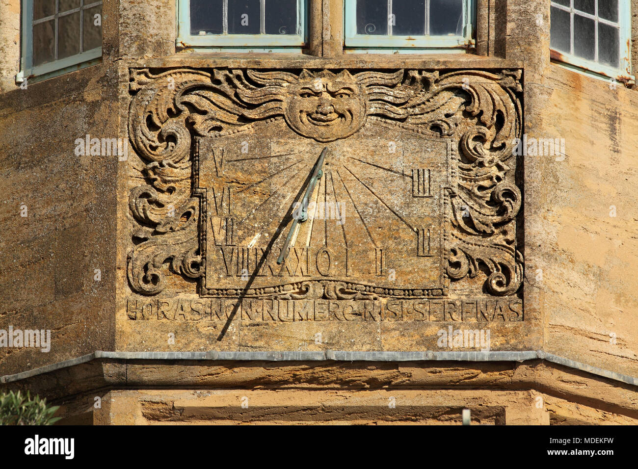 Un orologio solare su un muro di casa, con numeri romani, un immagine antropomorfica del sole e del moto vorticoso scrollwork, Lyme Regis, Dorset. Foto Stock