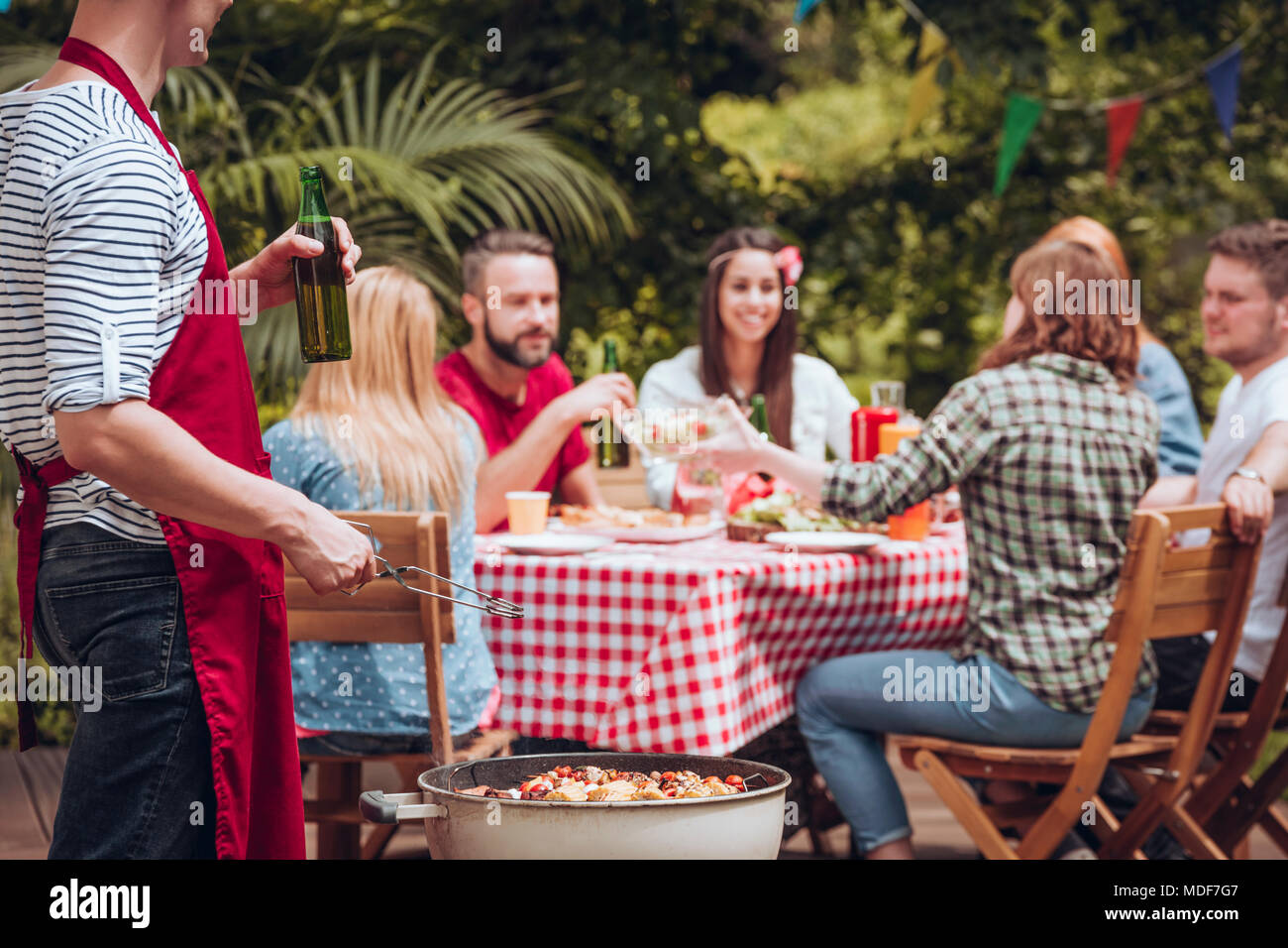 Uomo in un grembiule grigliare e bere birra mentre i suoi amici stanno mangiando in un tavolo in giardino Foto Stock