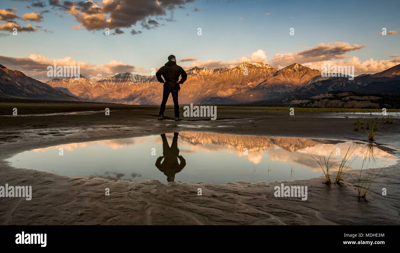 Un uomo sta con la sua riflessione in una piscina di acqua guardando le Saint Elias montagne al tramonto, Parco Nazionale Kluane e riserva Foto Stock