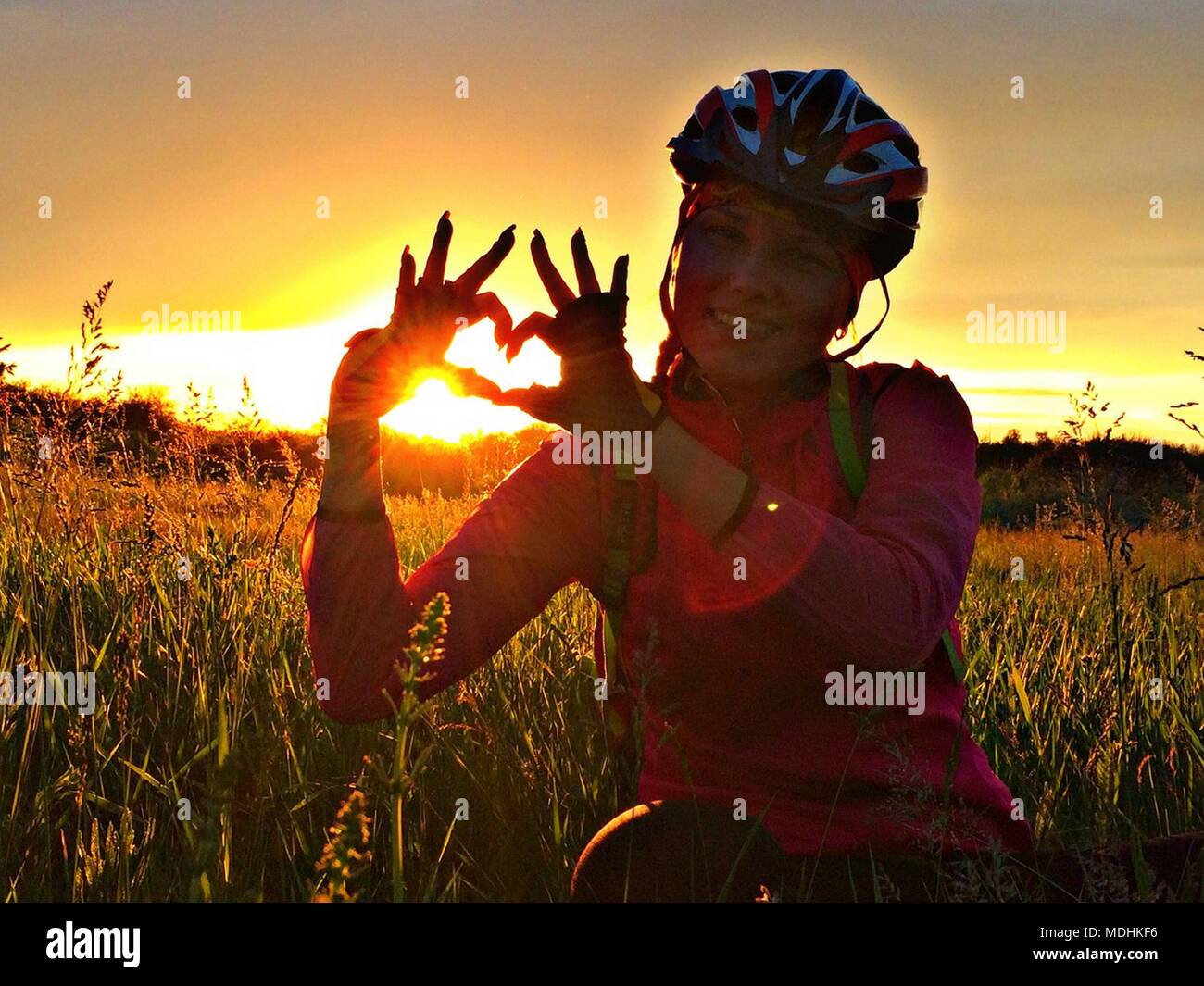 Ragazza in un casco per bicicletta sul campo mostra il simbolo del cuore. Le mani della donna che mostra forma di cuore con il tramonto del sole in background Foto Stock Ragazza in un casco per bicicletta sul campo mostra il simbolo del cuore. Le mani della donna che mostra forma di cuore con il tramonto del sole in background Foto Stock