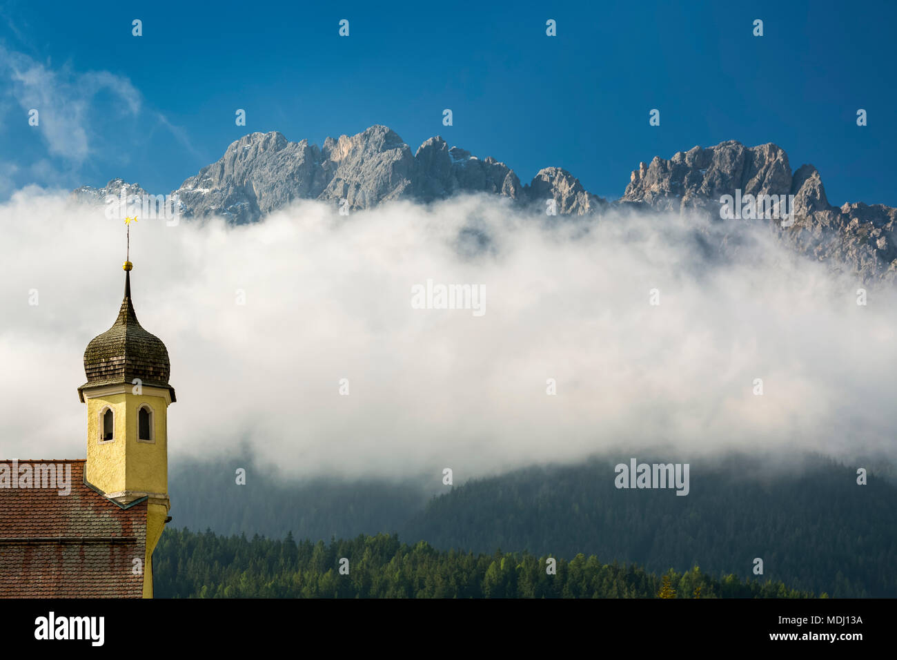 Guglia della chiesa nella valle con picchi di montagna del peering fuori dalle nuvole che coprono la montagna; San Candido, Bolzano, Italia Foto Stock