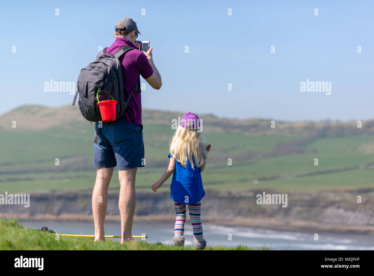 Un uomo e una ragazza bambino in piedi sulla cima di una scogliera che guarda al mare in abiti estivi. Egli è di scattare una foto con un telefono cellulare. Foto Stock