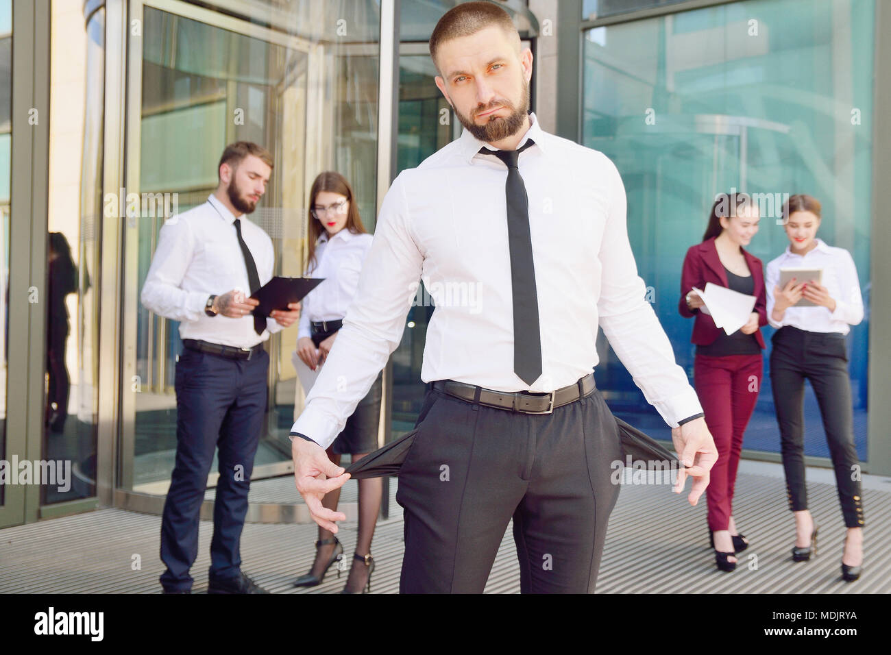 In bancarotta. Un giovane uomo in pantaloni neri e una camicia bianca  dimostra la sua vuota tasche in background di un gruppo di impiegati di  ufficio. Il licenziamento fro Foto stock -
