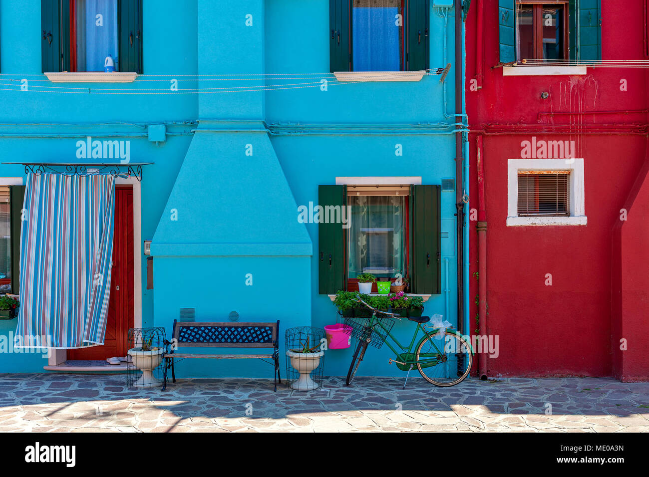 Piccola panca e bicicletta nella parte anteriore di piccole case dipinte di blu e di rosso a Burano, Italia. Foto Stock