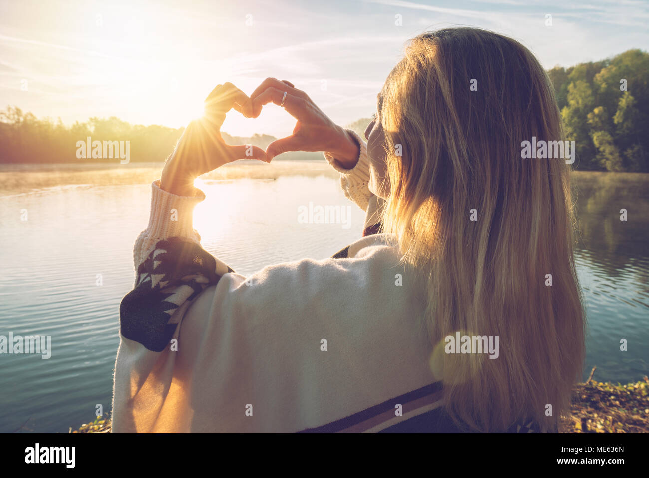 Giovane donna in piedi dal lago a sunrise facendo una forma di cuore dito sul telaio il bellissimo paesaggio, riflessione sull'acqua. La gente ama viaggiare Foto Stock