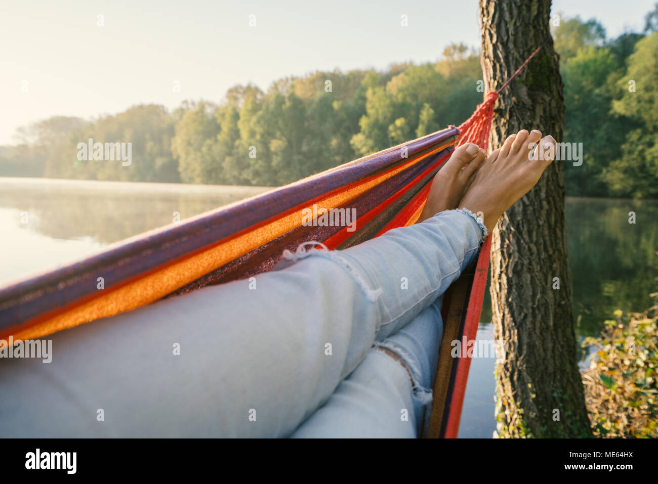 Punto di vista personale della donna relax su amaca in riva al lago nella stagione primaverile, scalzi, la luce del sole riflesso sulla superficie del lago. Pigrizia di viaggio Foto Stock