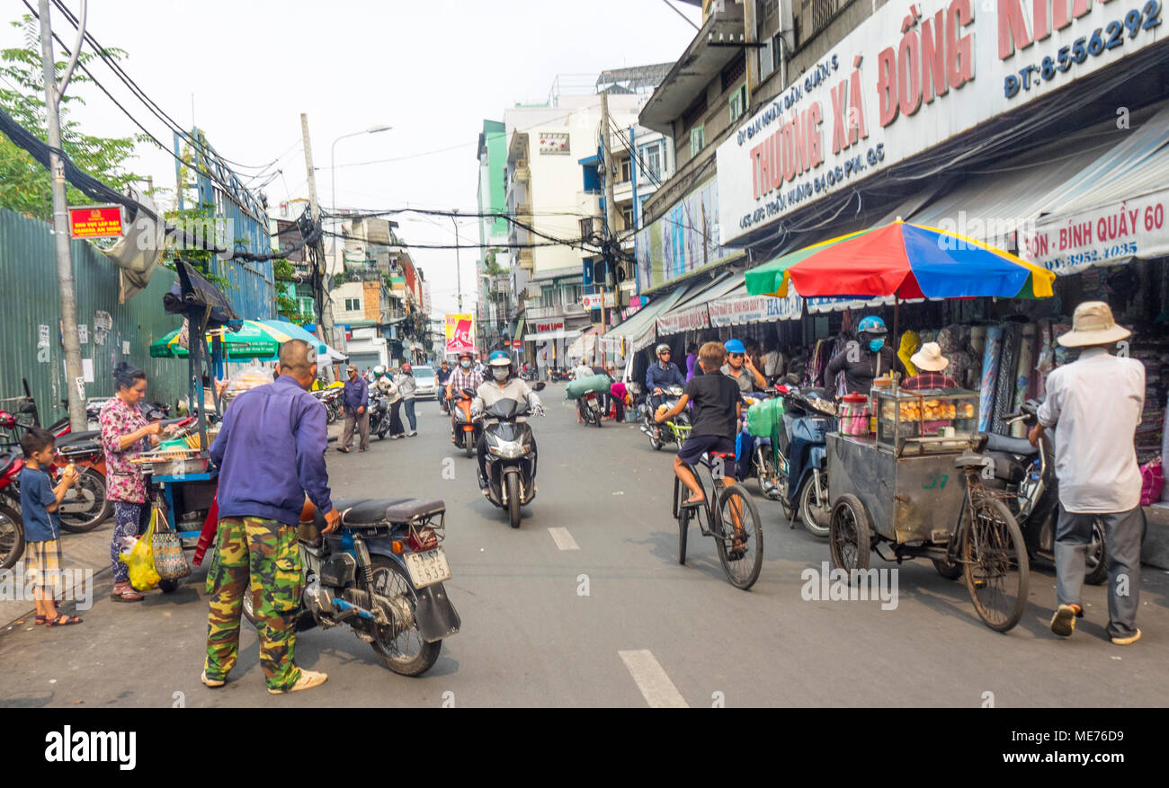 Motociclisti, ciclisti e ambulanti condividendo la strada di fronte a mercati di tessuto nella città di Ho Chi Minh, Vietnam. Foto Stock