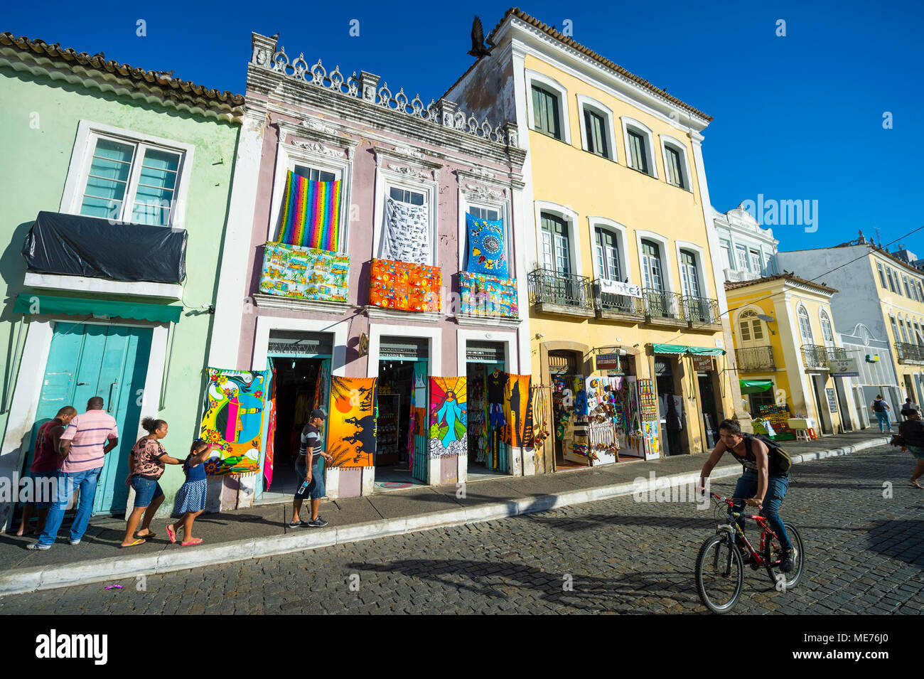SALVADOR, Brasile - circa Febbraio, 2018: pedoni e ciclisti passano davanti colorati edifici coloniali la presentazione e la vendita di luminoso negozio di souvenir Foto Stock