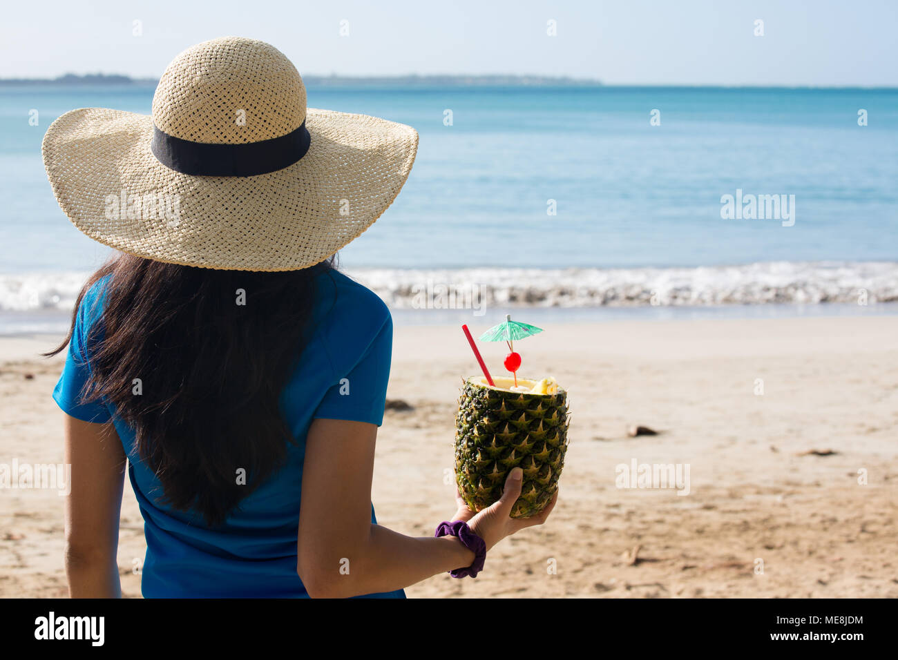Closeup ritratto, donna in maglietta blu e marrone cappello tenendo la pina colada rum ananas bevanda miscelata con paglia e piccolo ombrello, guardando fuori towa Foto Stock