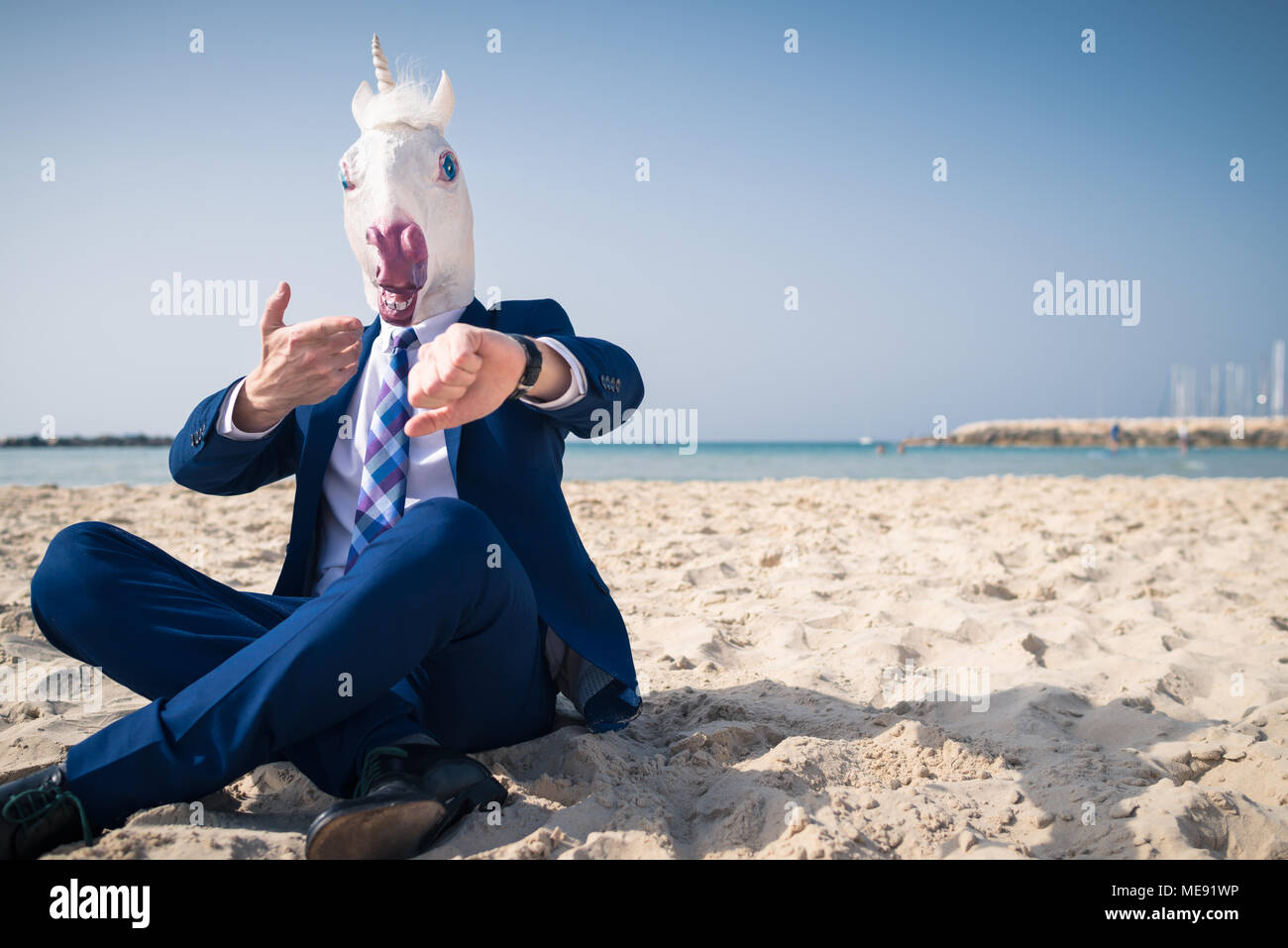 Elegante manager nella maschera di comico ed elegante abito siede sulla spiaggia e si affaccia sull'orologio. Funny Man gesticulating attivo sullo sfondo del mare e del cielo Foto Stock