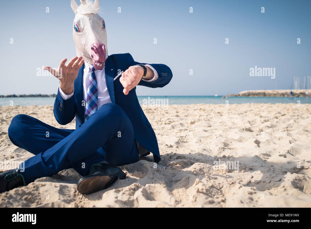 Elegante uomo in maschera divertente ed elegante abito siede sulla spiaggia e si affaccia sull'orologio. Paura l'uomo perse nel tempo ed è in ritardo per il business Foto Stock