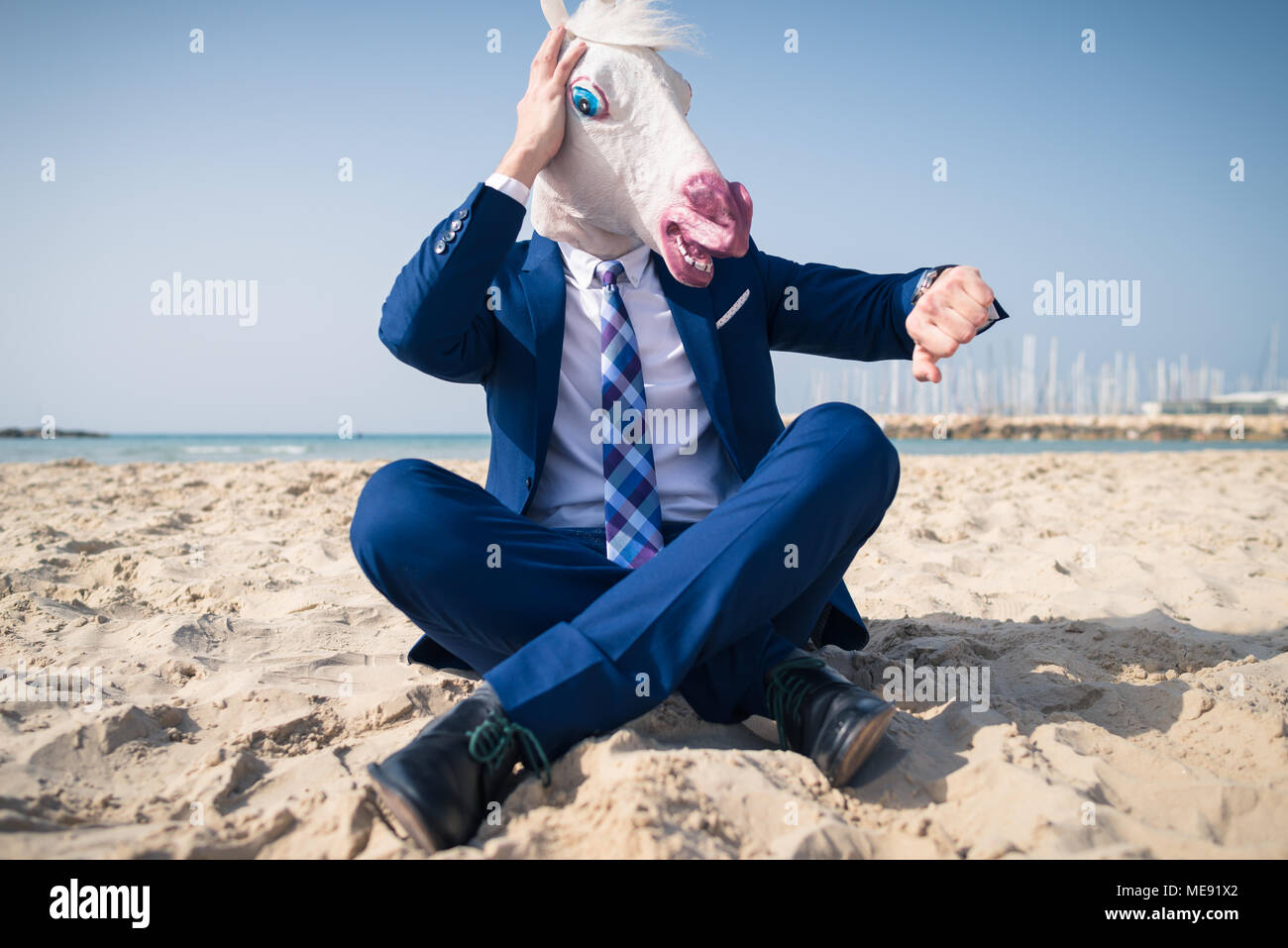 Elegante manager nella maschera di comico ed elegante abito siede sulla spiaggia e si affaccia sull'orologio. Paura l'uomo perse nel tempo ed è in ritardo per il business Foto Stock