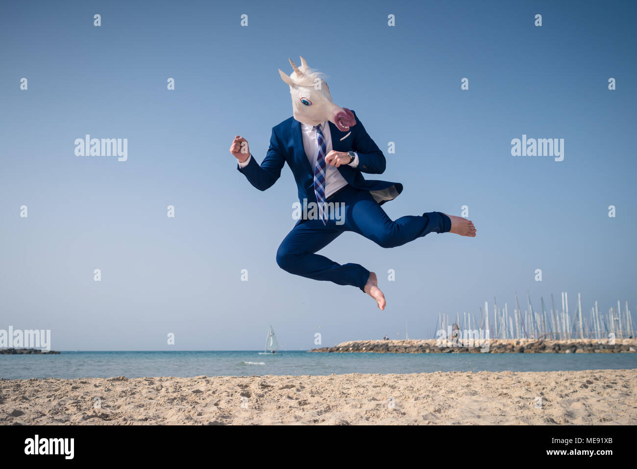 Elegante manager nella maschera divertente ed elegante abito salta sulla spiaggia. Grave fighter colpisce in volo. Unicorn sullo sfondo del mare e del cielo Foto Stock