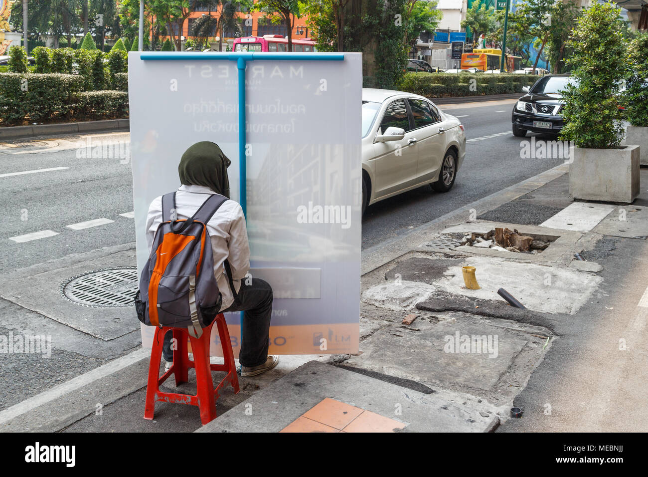 Uomo seduto su una sedia in plastica dal lato della Sukhumvit Road tenendo su un pannello pubblicitario al traffico passante, Bangkok, Thailandia Foto Stock