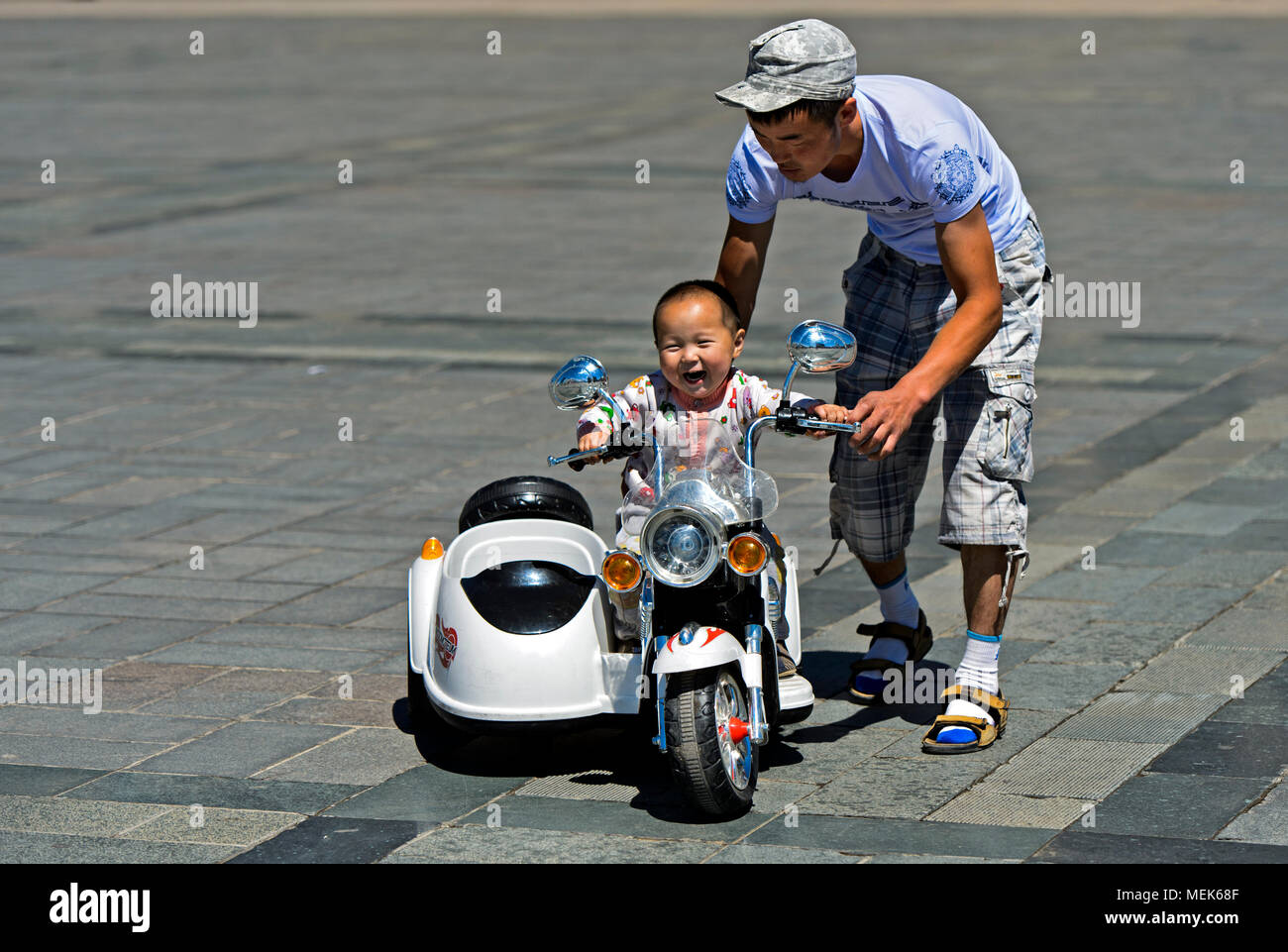 Il Toddler boy cavalcare un giocattolo elettrico motore-bike con la guida di suo padre sulla piazza Sukhbaatar, Ulaanbaatar, in Mongolia Foto Stock