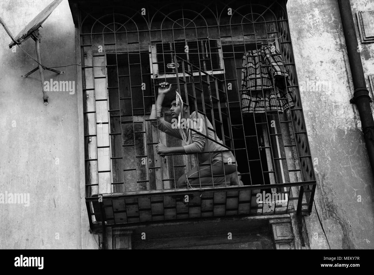 Giovane uomo su un balcone a gabbia, Margao, Goa, India. Egli sembra in qualche modo la speranza come se sognare un futuro migliore guardando fuori dal di dentro il suo ambiente circostante. Foto Stock