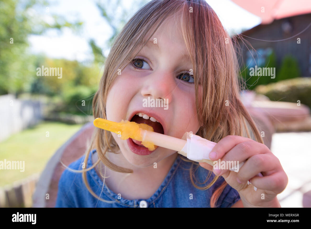 Ritratto di close-up di faccia di quattro anni ragazza bionda mangiare arancione o giallo per lecca-lecca di ghiaccio o ghiaccioli Foto Stock
