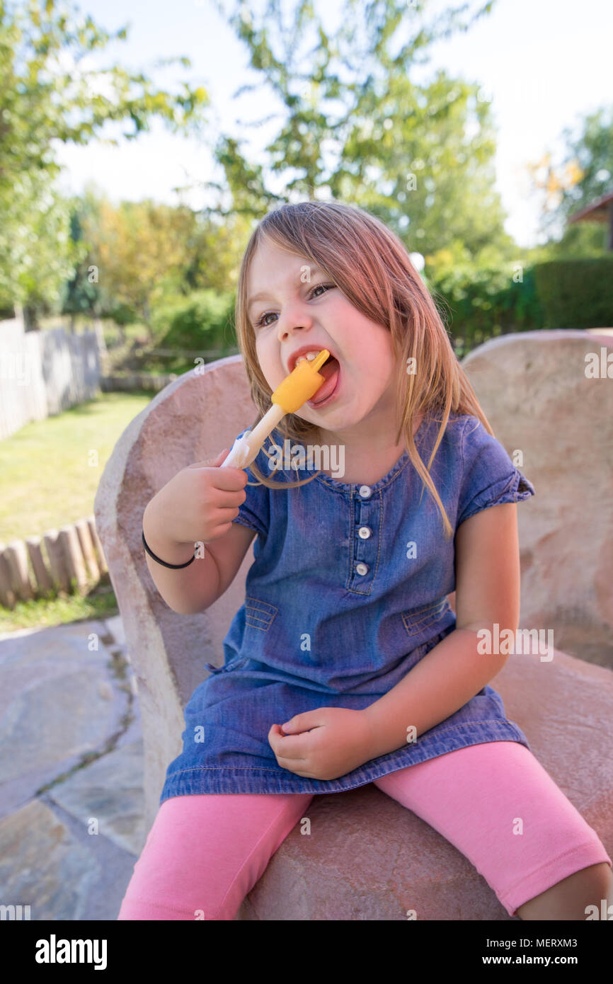 Quattro anni ragazza bionda con blu denim dress seduta nel parco pubblico, mordere arancione o giallo per lecca-lecca di ghiaccio o ghiaccioli Foto Stock