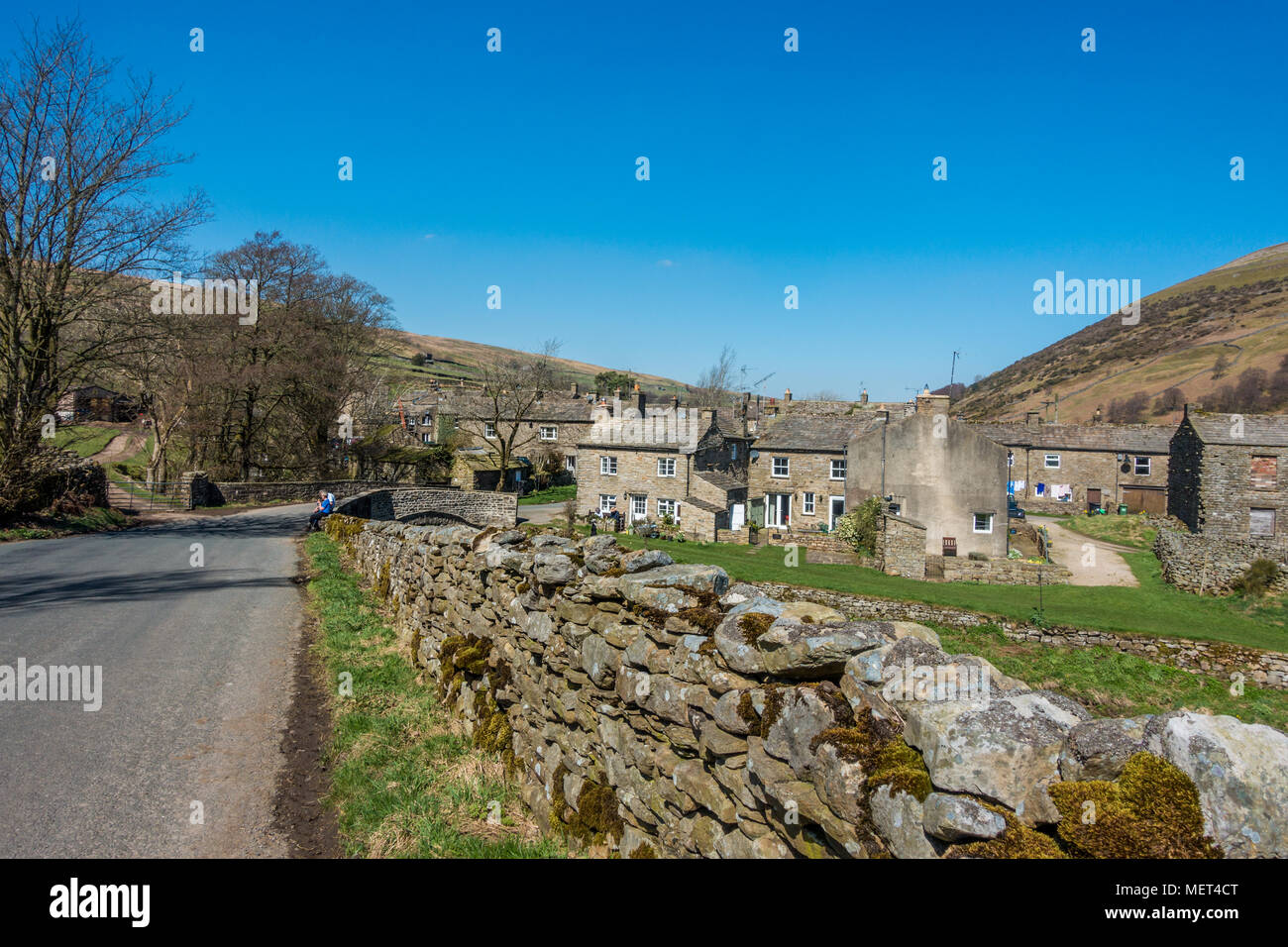 Le pittoresche case in pietra e il ponte nel villaggio di Thwaite su una bella giornata con due escursionisti, Yorkshire Dales, REGNO UNITO Foto Stock