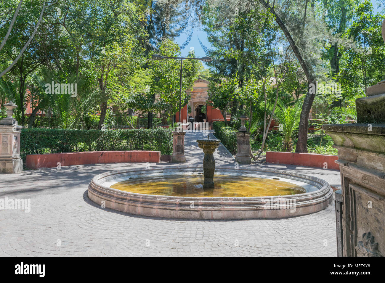 Vista serena di Benito Juarez Parco con alberi, pietra fontana, colonne di pietra e il sentiero in pietra, in una giornata di sole in San Miguel De Allende, Messico Foto Stock