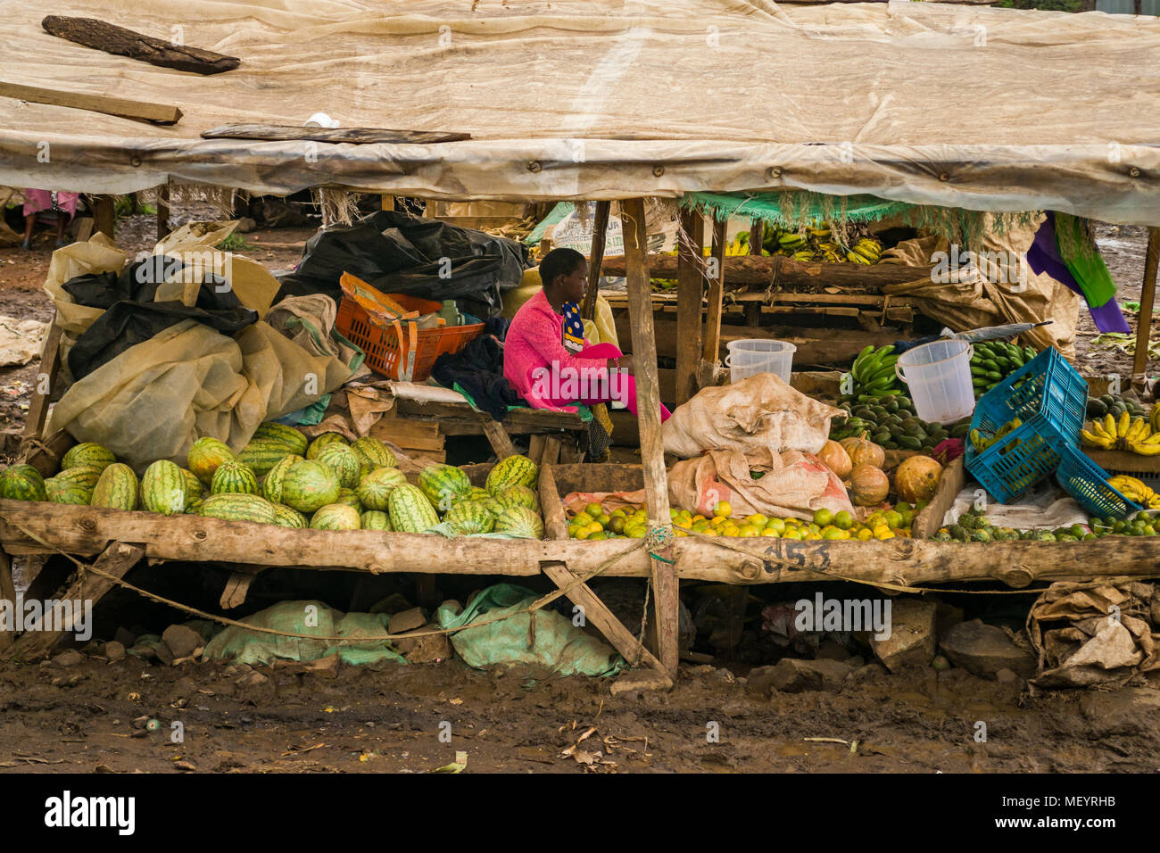 Una ragazza si siede sotto la copertura di un mercato in stallo con cocomero, limone e banane sul display per la vendita, Africa orientale Foto Stock
