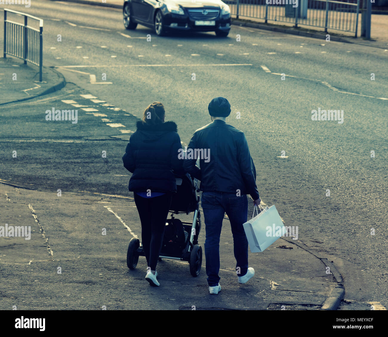 La popolazione locale coppia giovane uomo e donna shopping spingendo la PRAM su strada marciapiede marciapiede del traffico su strada attraversando Castlemilk, Glasgow, Regno Unito Foto Stock