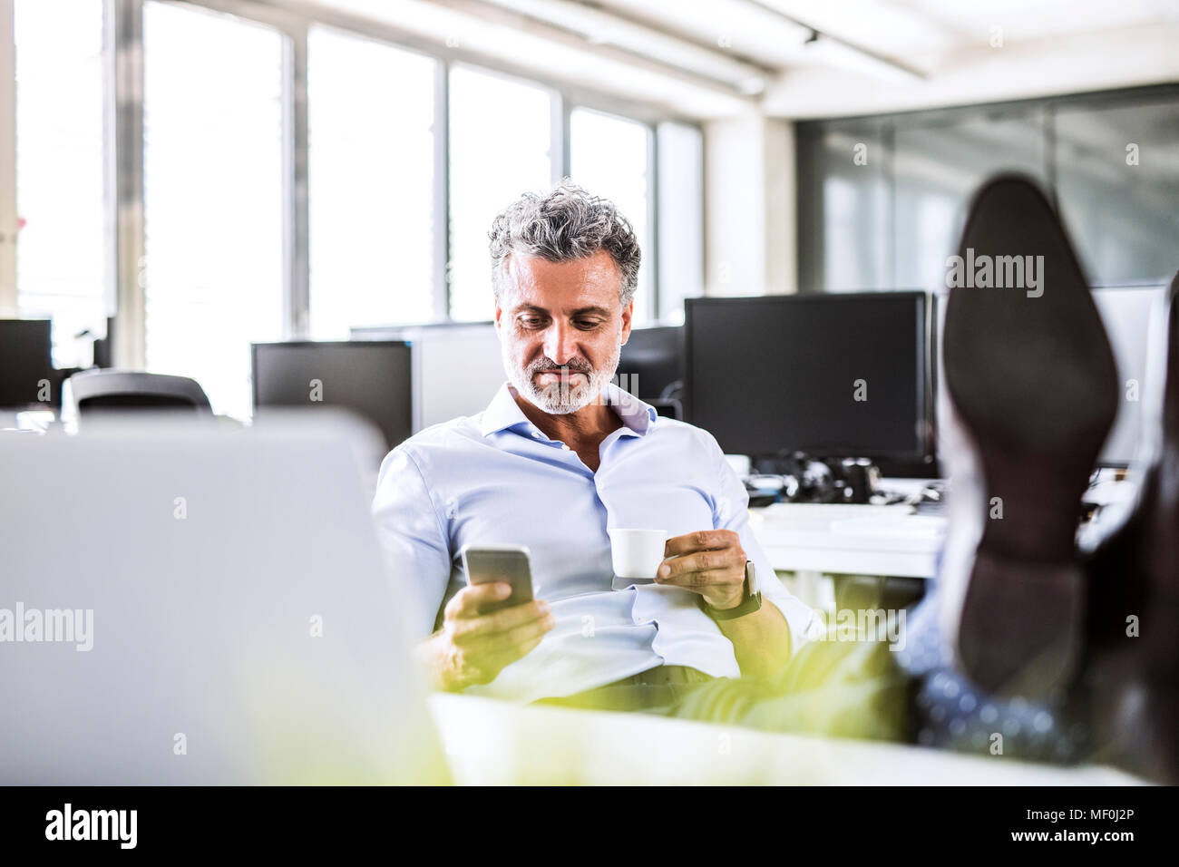 Rilassata imprenditore maturo seduto alla scrivania in ufficio tramite telefono cellulare e di bere il caffè Foto Stock