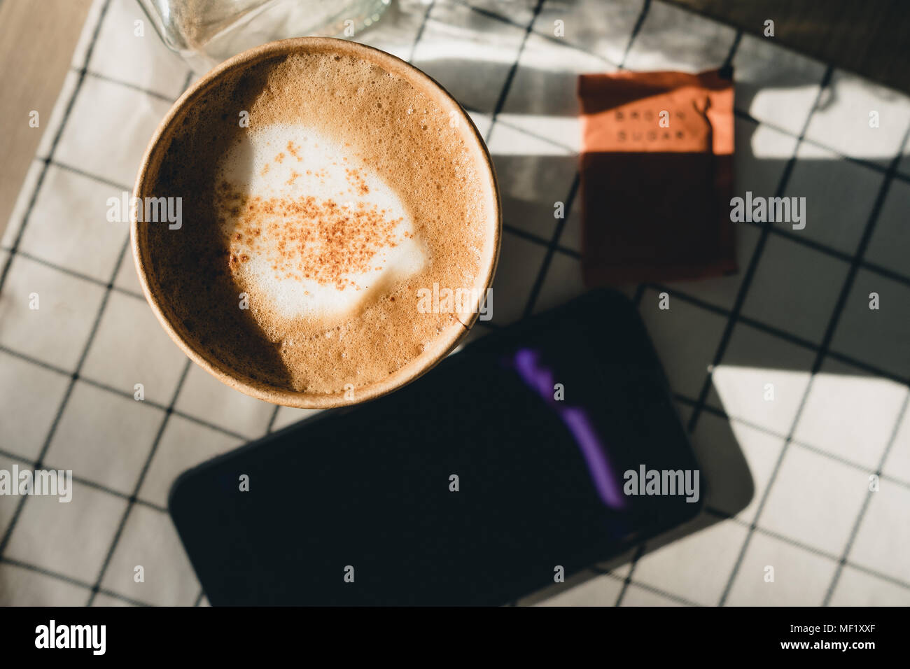 Cappuccino in take away tazza da caffè con telefono cellulare sul panno tavolo con fiori secchi,gli occhiali da sole sul tavolo di legno con la luce del sole ombra rigido in corrispondenza della finestra in ev Foto Stock