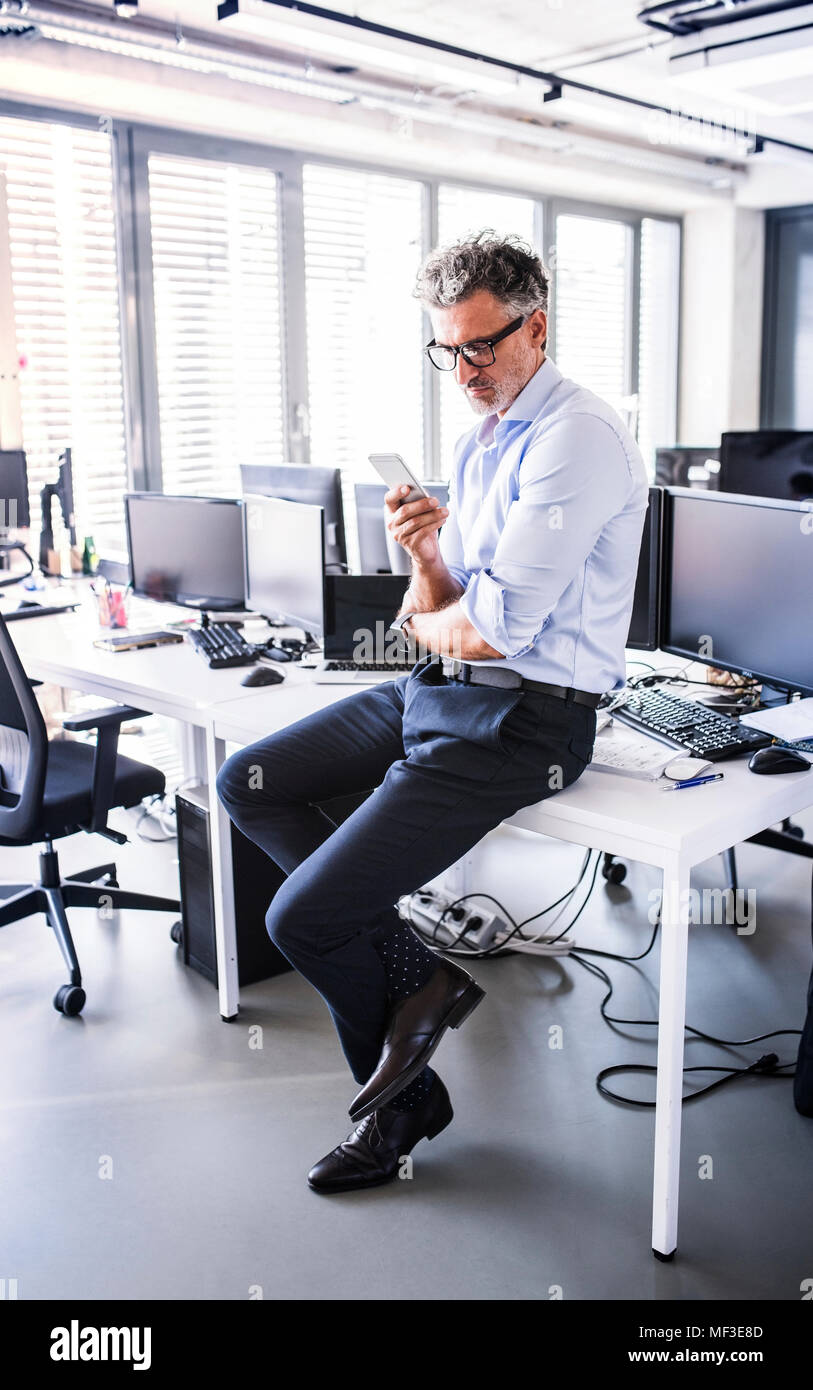 Imprenditore maturo seduto sulla scrivania in ufficio utilizza lo smartphone Foto Stock