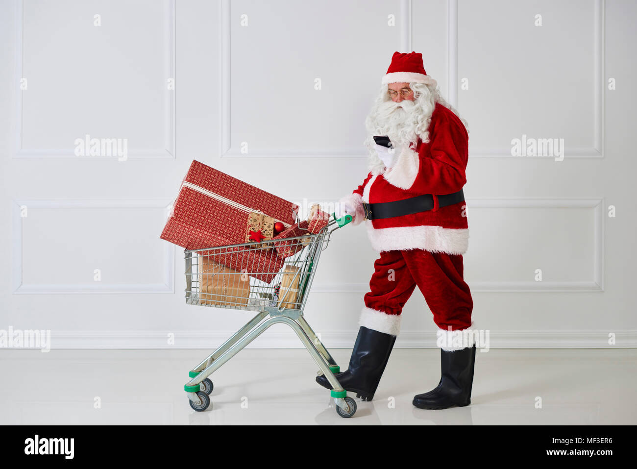 Babbo Natale che porta i regali di Natale in un carrello di shopping mentre guarda il telefono cellulare Foto Stock