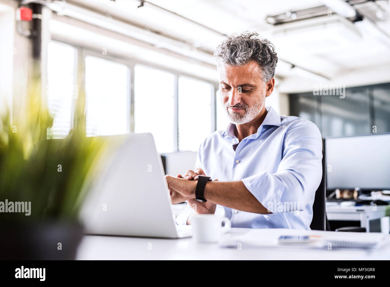 Imprenditore maturo seduto alla scrivania in ufficio guardando smartwatch Foto Stock