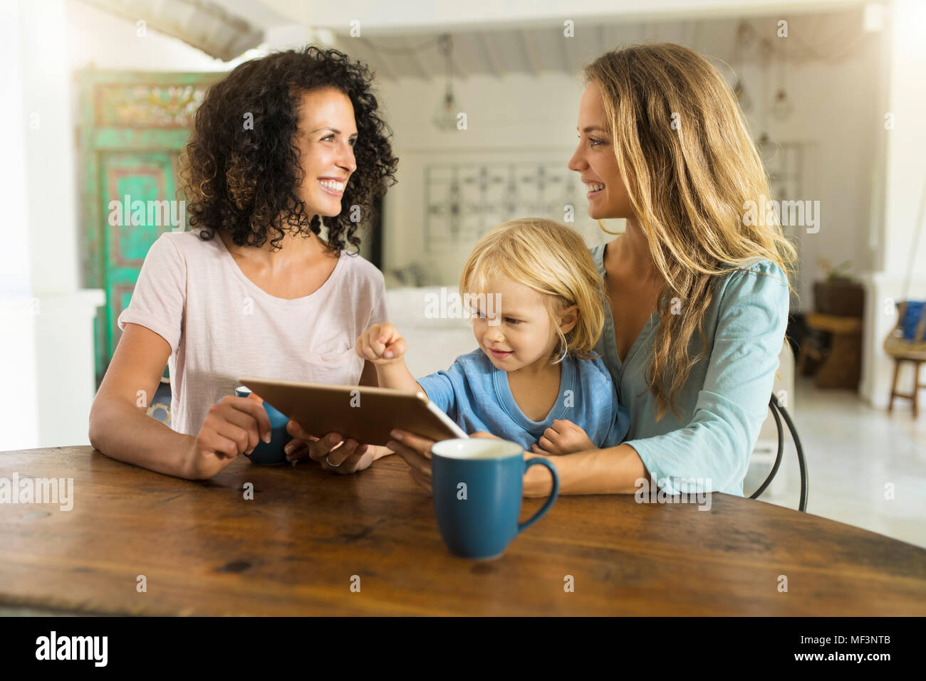 Due donne sorridenti con un bambino utilizzando tablet al tavolo della cucina Foto Stock