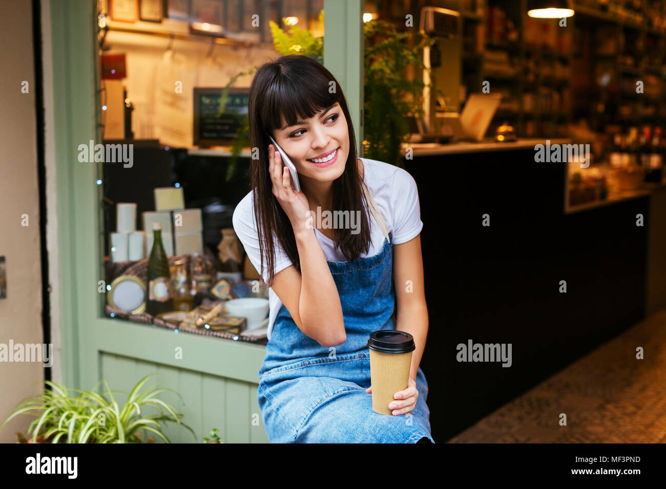 Donna sorridente seduto alla porta di ingresso di un negozio parlando al cellulare Foto Stock