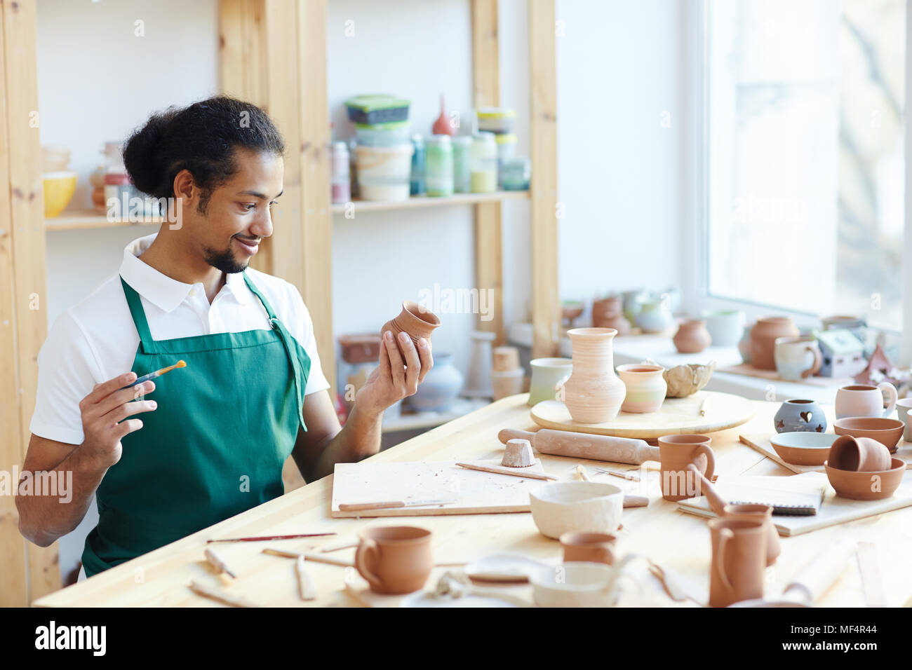 Giovani razza mista potter maschile seduto al tavolo del workshop, l'applicazione di smalto sulla artigianale di pentola di creta con pennello e sorridente Foto Stock